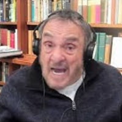 He is talking while wearing headphones with his book collection in the background.